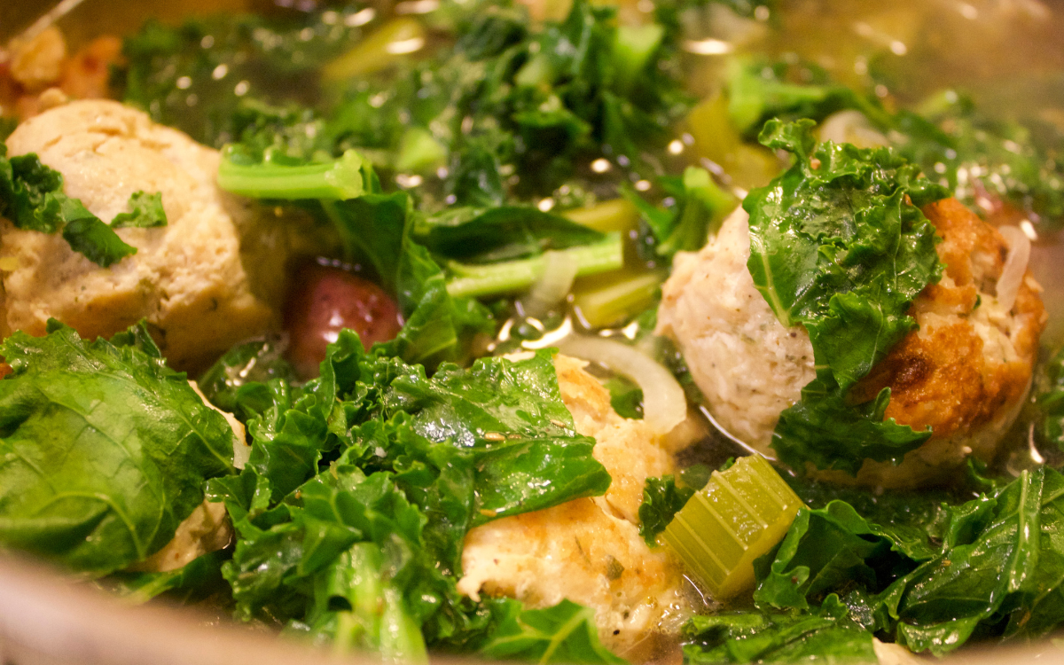 A close up of bright green, fresh kale, celery and turkey meatballs in a soup broth pot.