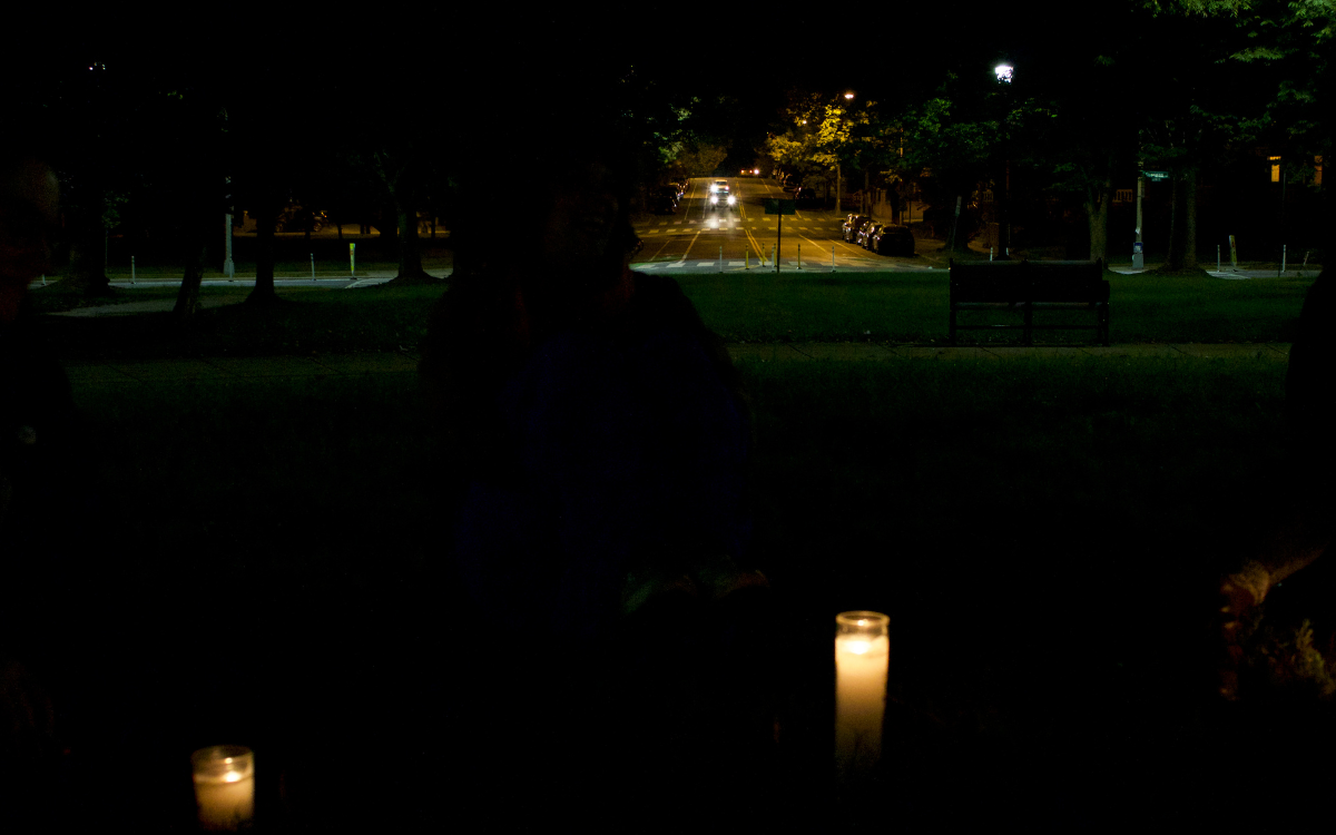 A shadowed silhoutette of a person sitting in a park illuminated by candles in front of them and a street illuminated by car headlights behind them.