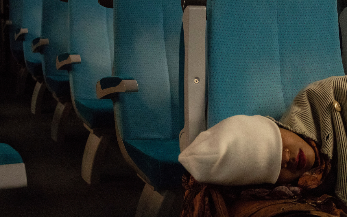 A Black woman wearing a white beanine cap to cover her eyes naps on a train with blue cushioned chairs.