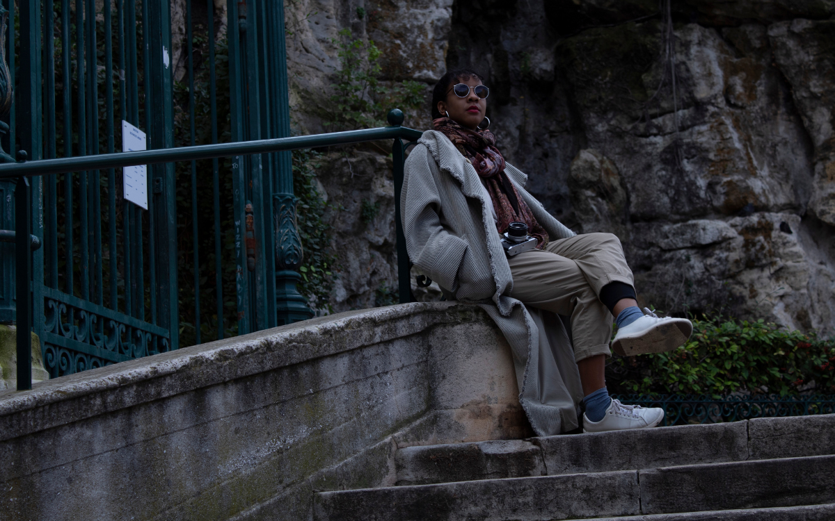 A Black woman in a long coat, hoop earrings and sunglasses with a film camera on her person sits on a railing ledge over steps in Marseille, France.